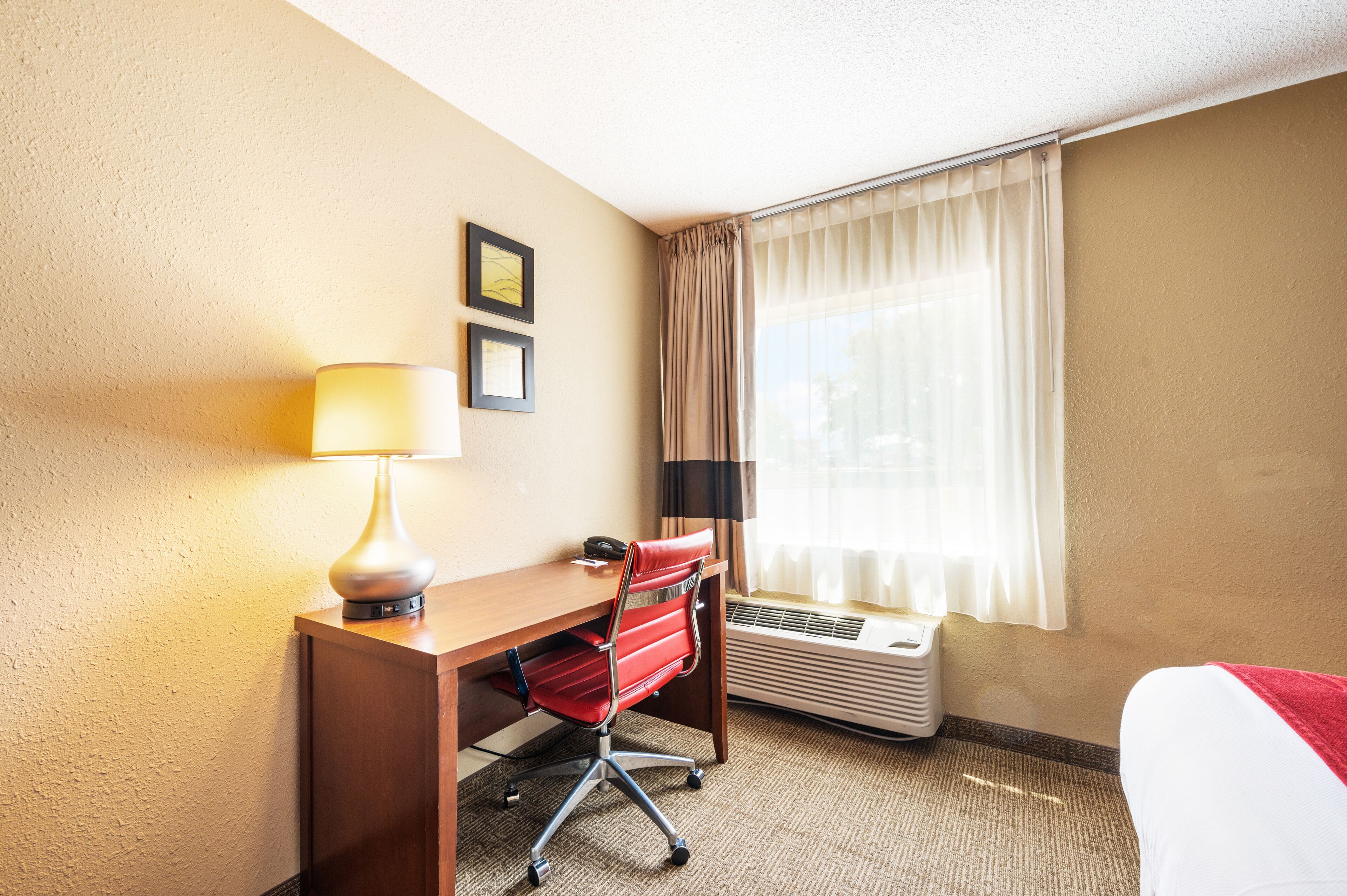 A hotel room with a desk, chair, lamp, air conditioning unit, and a bed with a red blanket.