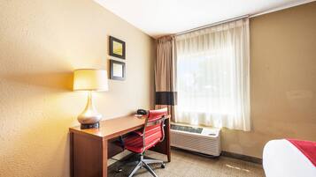 A hotel room with a desk, chair, lamp, air conditioning unit, and a bed with a red blanket.