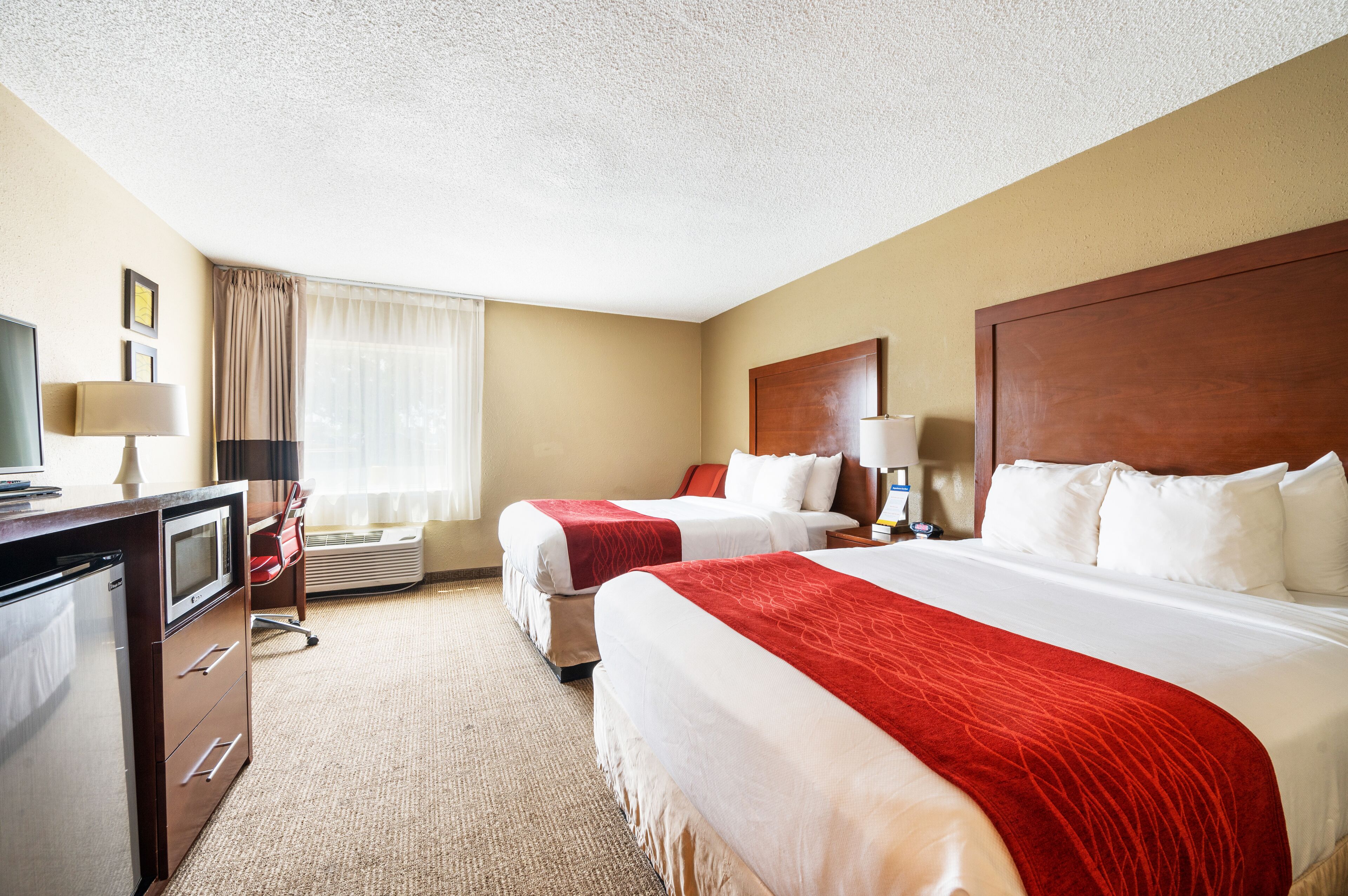 A hotel room with two beds, a desk with a computer, a lamp, and a window with curtains.