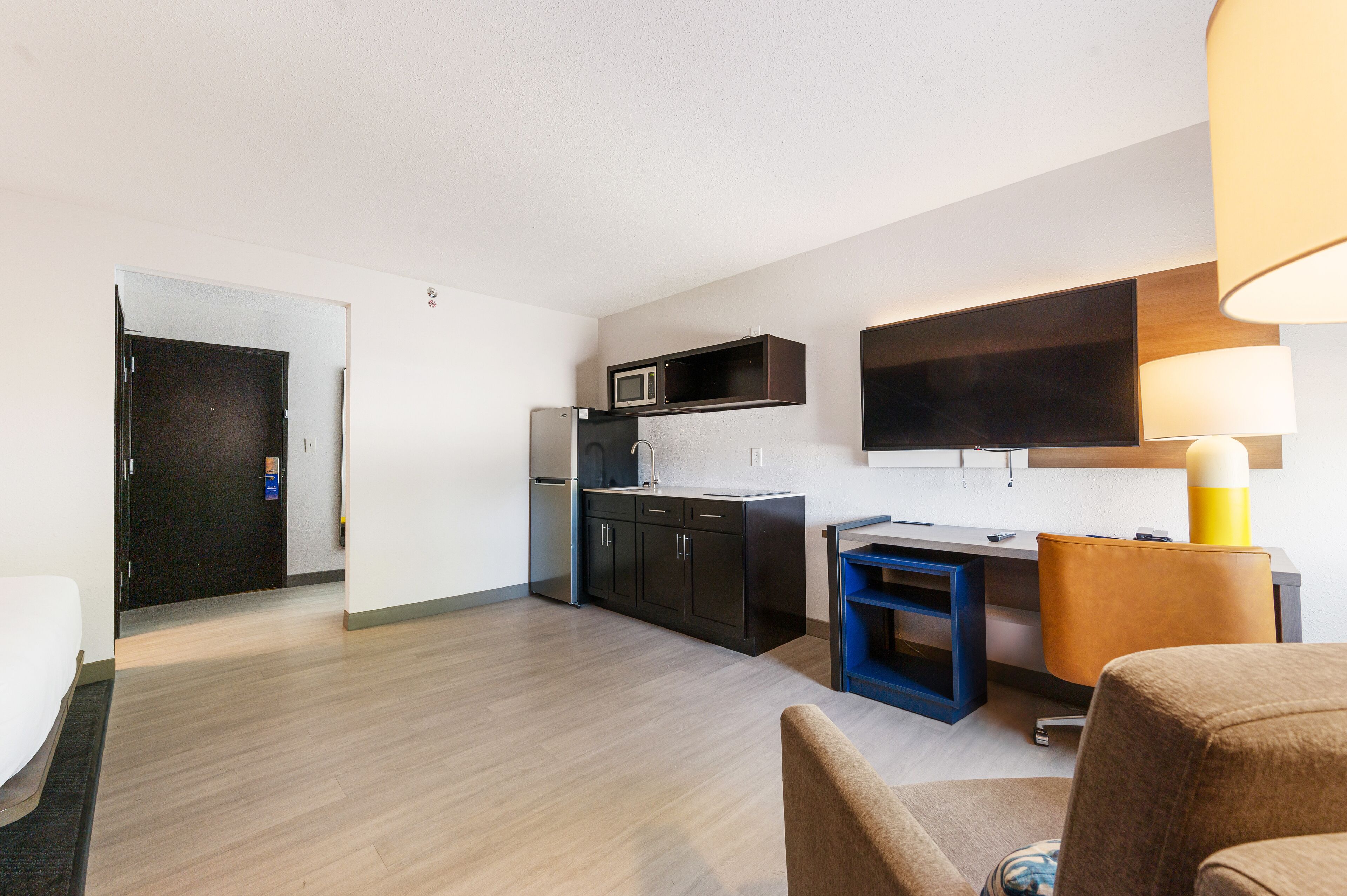 Una habitación de hotel moderna con kitchenette, un televisor de pantalla plana montado en la pared, un escritorio pequeño con una silla y un sofá.