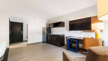Una habitación de hotel moderna con kitchenette, un televisor de pantalla plana montado en la pared, un escritorio pequeño con una silla y un sofá.