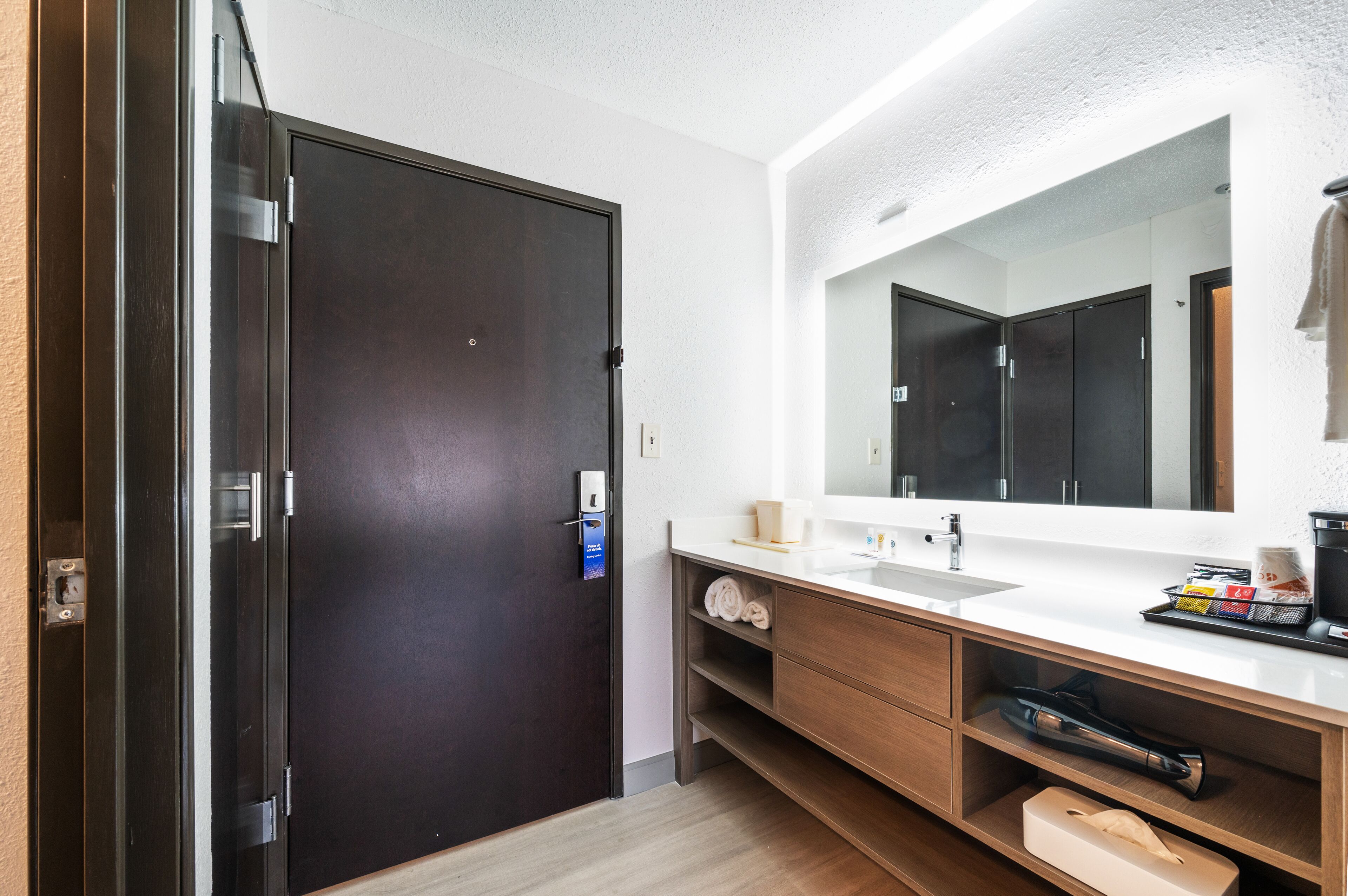 A modern bathroom with a dark door, a large mirror, a white sink, and wooden cabinets.