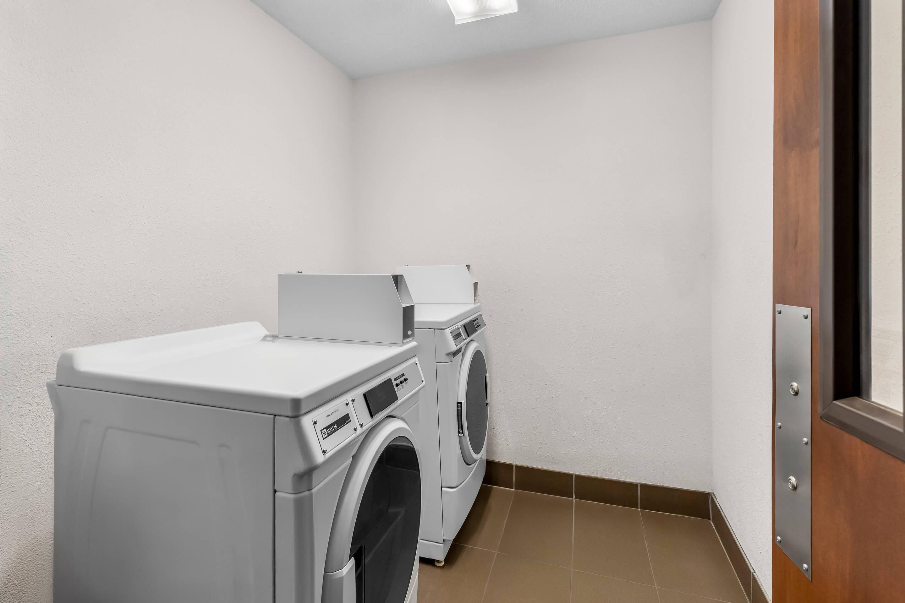 A laundry room with two front-loading washing machines and a dryer, white walls, and a wooden door.