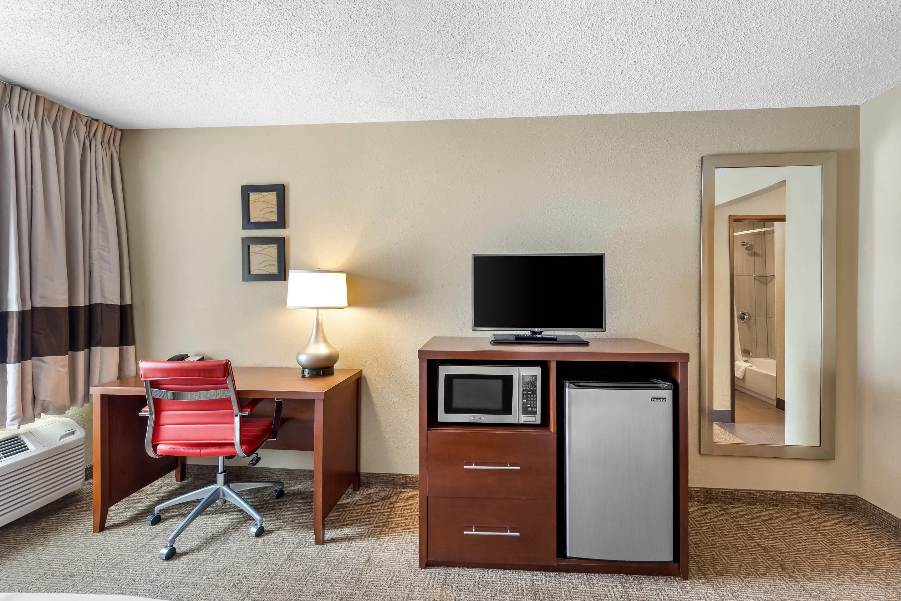 A hotel room with a desk, a chair, a television, a microwave, a mini-fridge, a lamp, and a mirror.