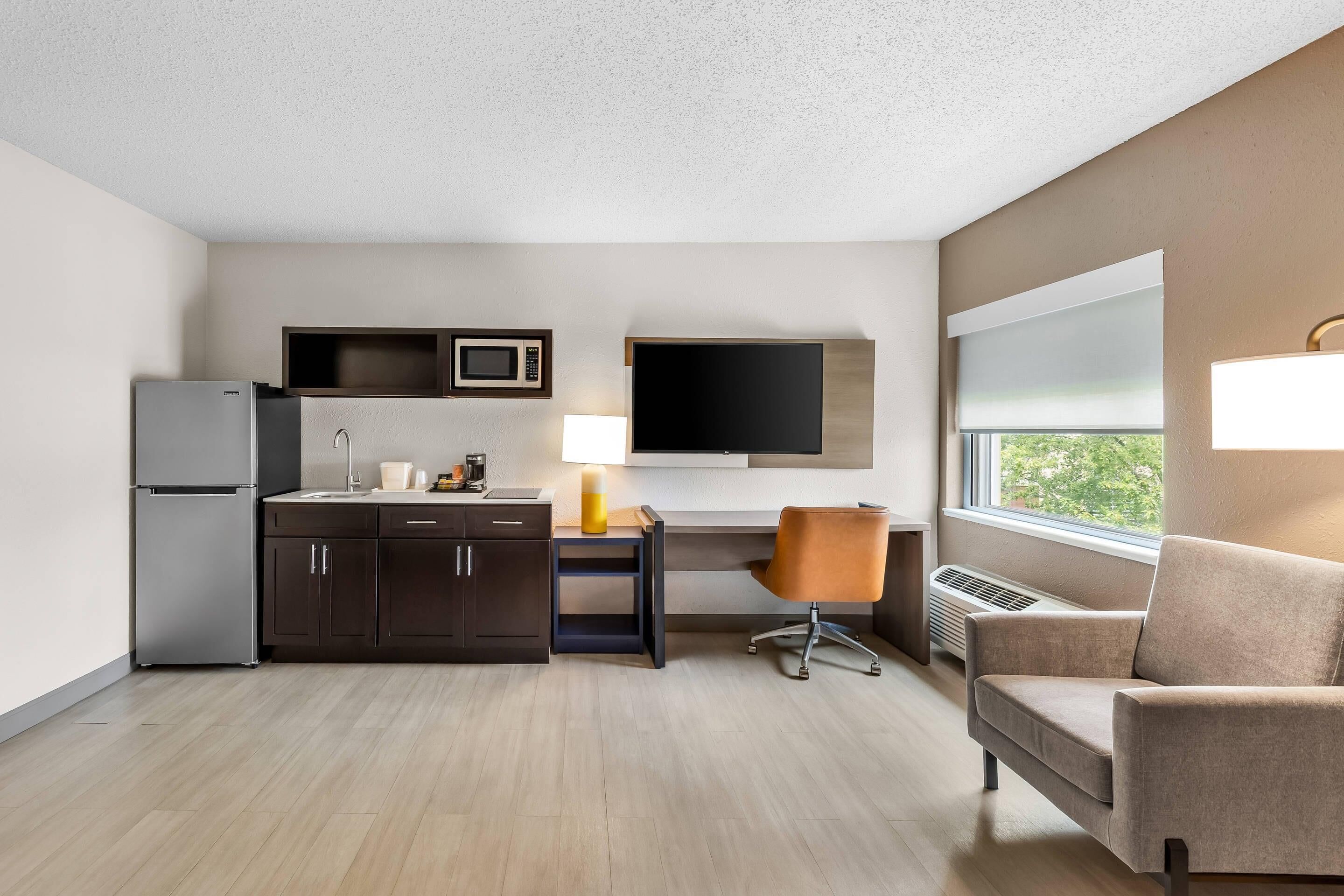 A compact hotel room with a kitchenette, a desk with a chair, a large window, and a flat-screen TV mounted on the wall.