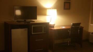 A hotel room with a desk, a television, a refrigerator, a chair, and a lamp.