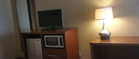 A hotel room with a bed, a desk with a lamp, a microwave, and a television on a cabinet.