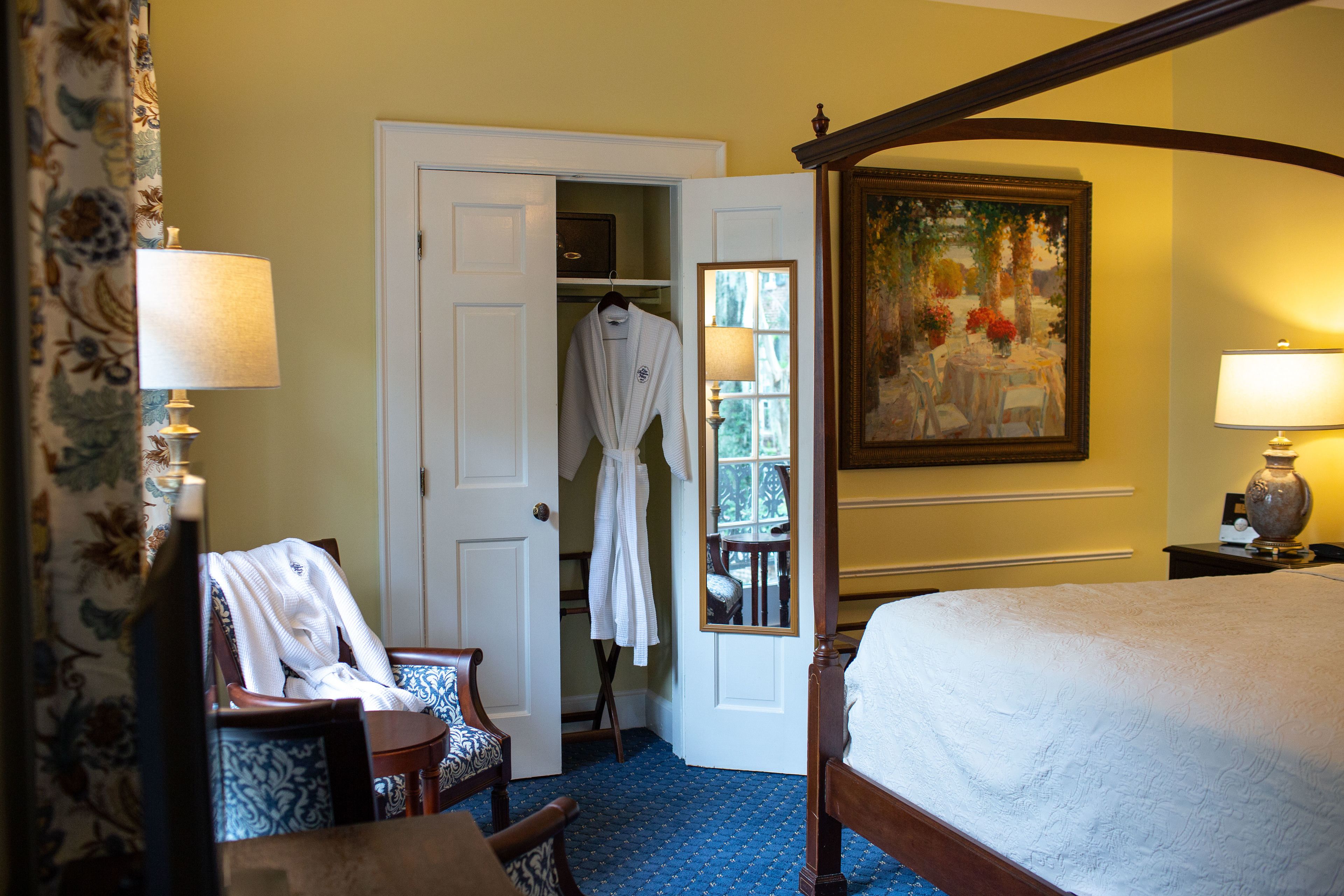 A hotel room with a bed, a chair, a robe hanging on a door, a painting on the wall, and a lamp on a nightstand.