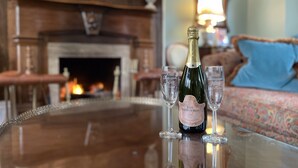 Uma garrafa de Los Monteros Cava Brut sobre uma mesa de vidro com quatro taças de vinho, uma lareira e um sofá com uma almofada estampada ao fundo.