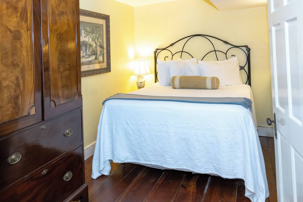 A neatly made bed with a white bedspread and pillows, a wooden dresser with drawers, and a framed picture on the wall.