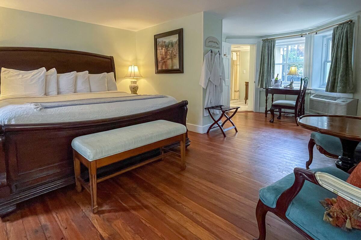 A hotel room with a large bed, a bedside table with a lamp, a chair, a small table, and a view of the outdoors through a window.
