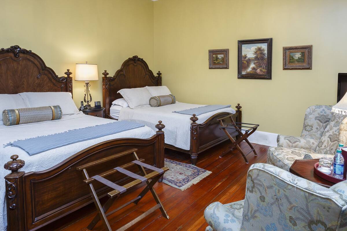 A double bed with a wooden headboard, a bedside table with a lamp, a chair with a tray, and a small table with a bottle and a cup.