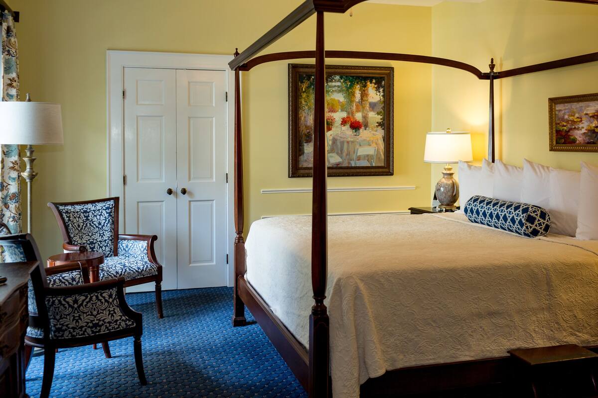 A four-poster bed with a white bedspread, a nightstand with a lamp, a chair with a patterned upholstery, and a door with a white frame.