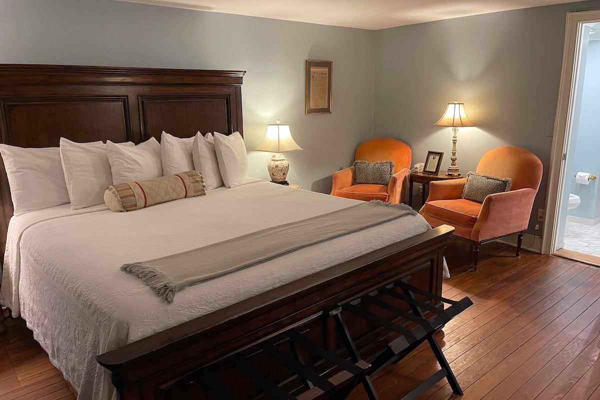 A neatly arranged bedroom with a large bed, two chairs, a wooden bench, and a framed certificate on the wall.