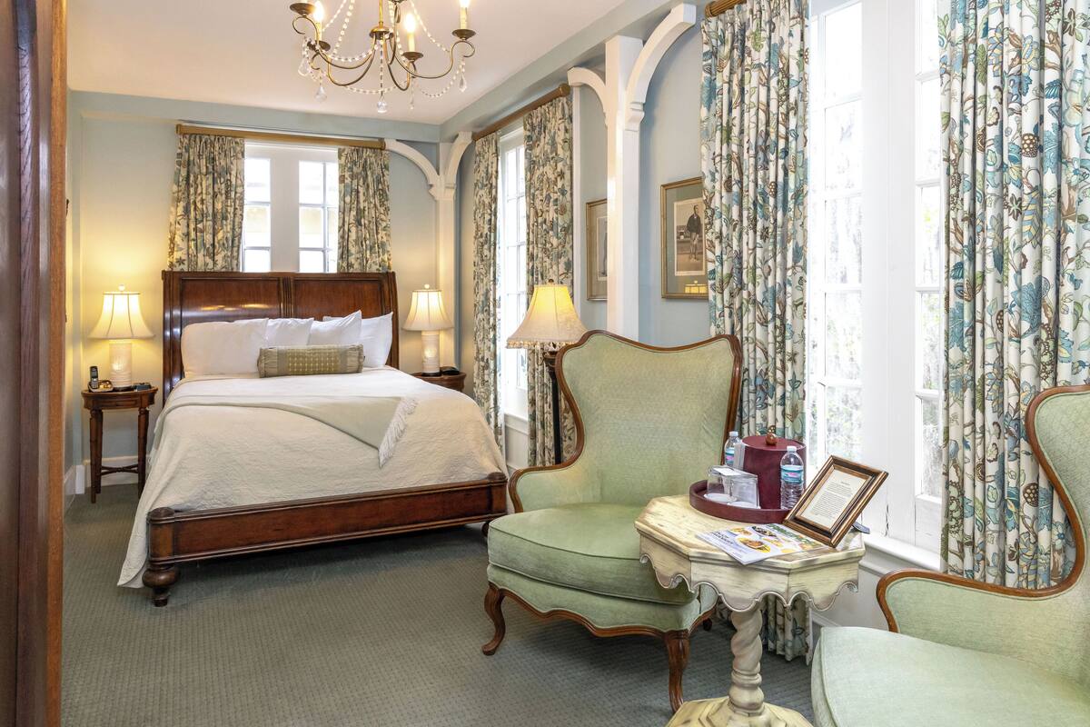 A hotel room with a large bed, a green armchair, a small table with a lamp, and floral curtains.