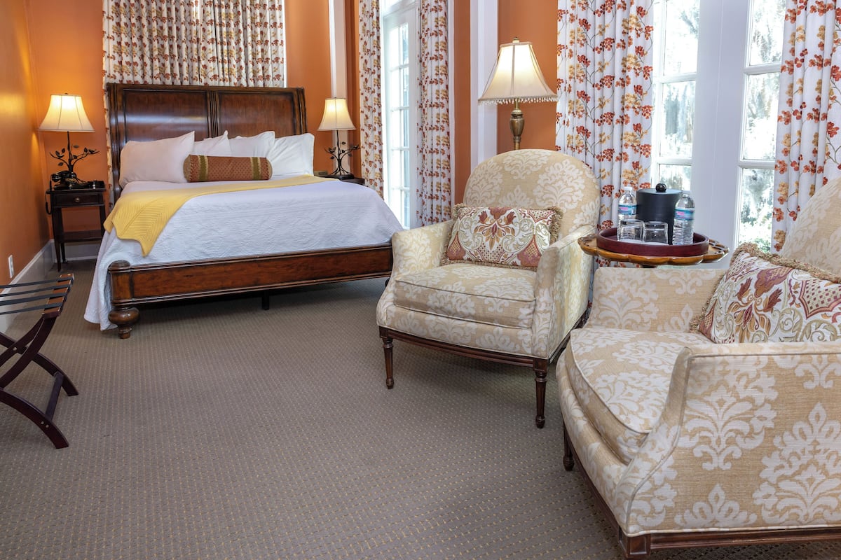 A hotel room with a large bed, two armchairs, a wooden nightstand with lamps, and a window with curtains.