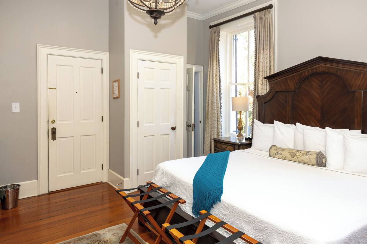 A bedroom with a large bed, a wooden headboard, a window with curtains, and a chandelier.