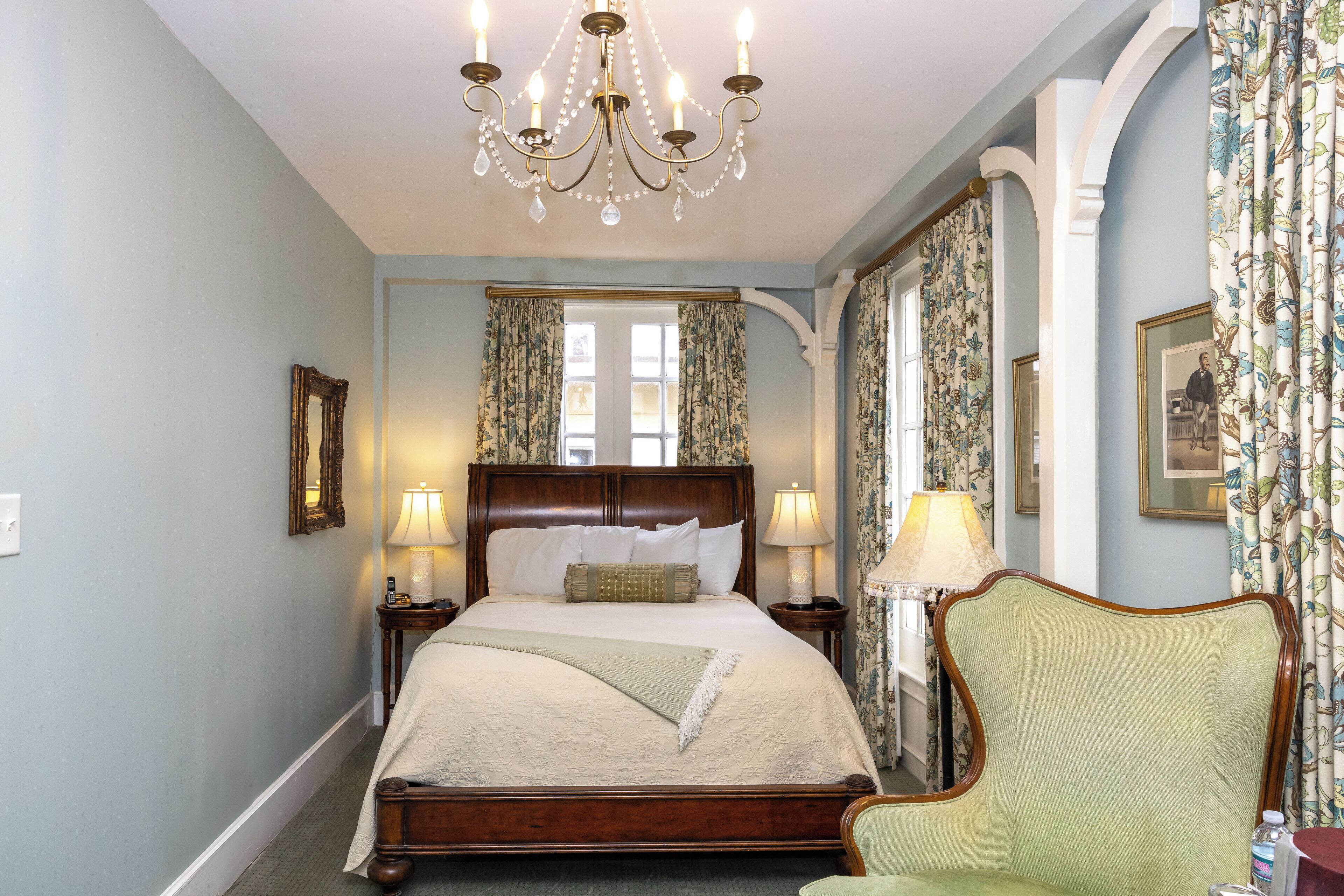 A bedroom with a large bed, two bedside tables, a chandelier, floral curtains, and a mirror on the wall.