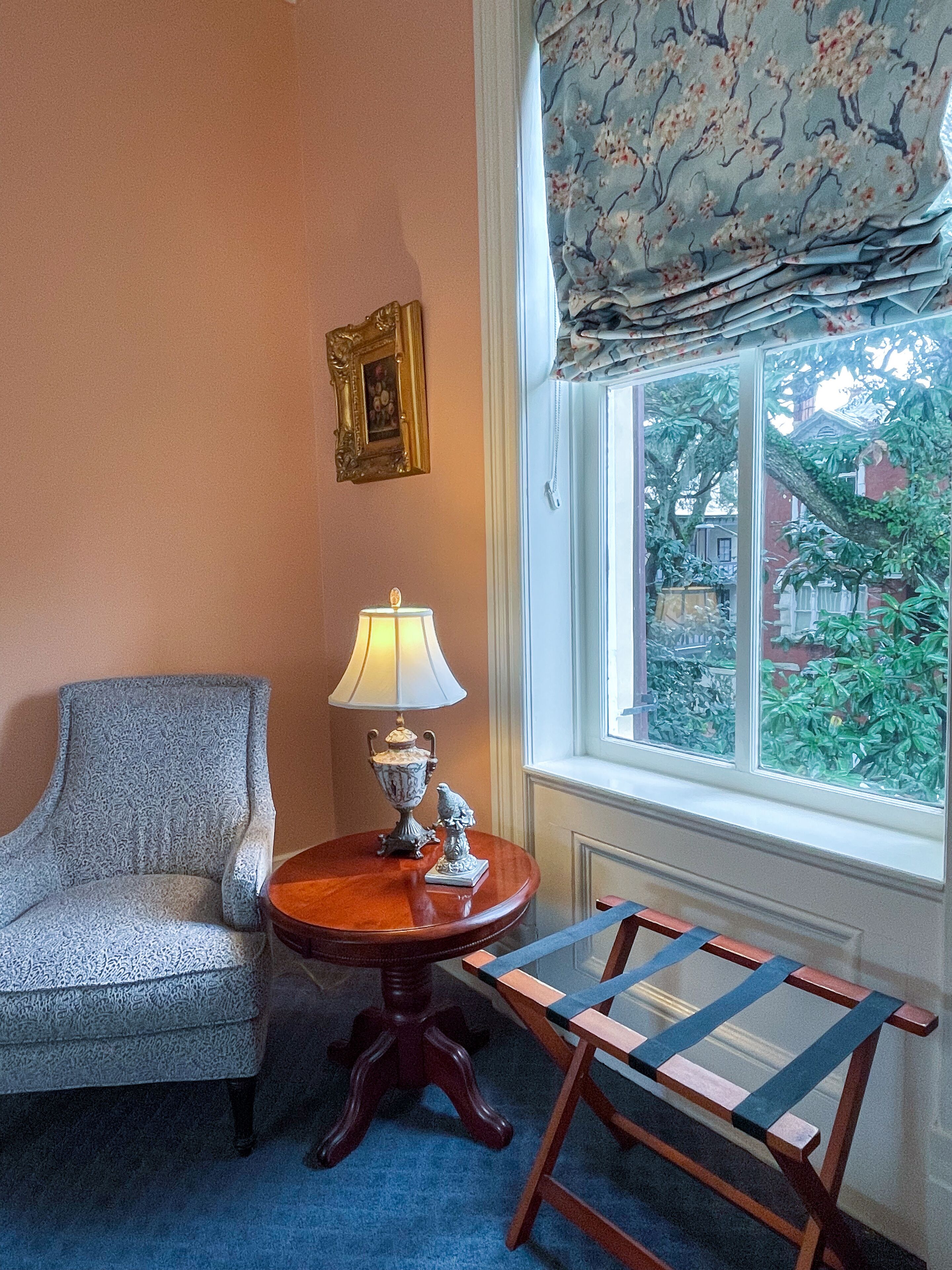 Una habitación con un sillón azul, una mesita de madera con una lámpara, y vistas a zonas verdes a través de una ventana con cortina floral.