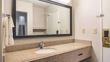 Baño con encimera de granito, lavamanos, espejo y una ducha visible en el reflejo.