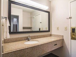 Baño con encimera de granito, lavamanos, espejo y una ducha visible en el reflejo.