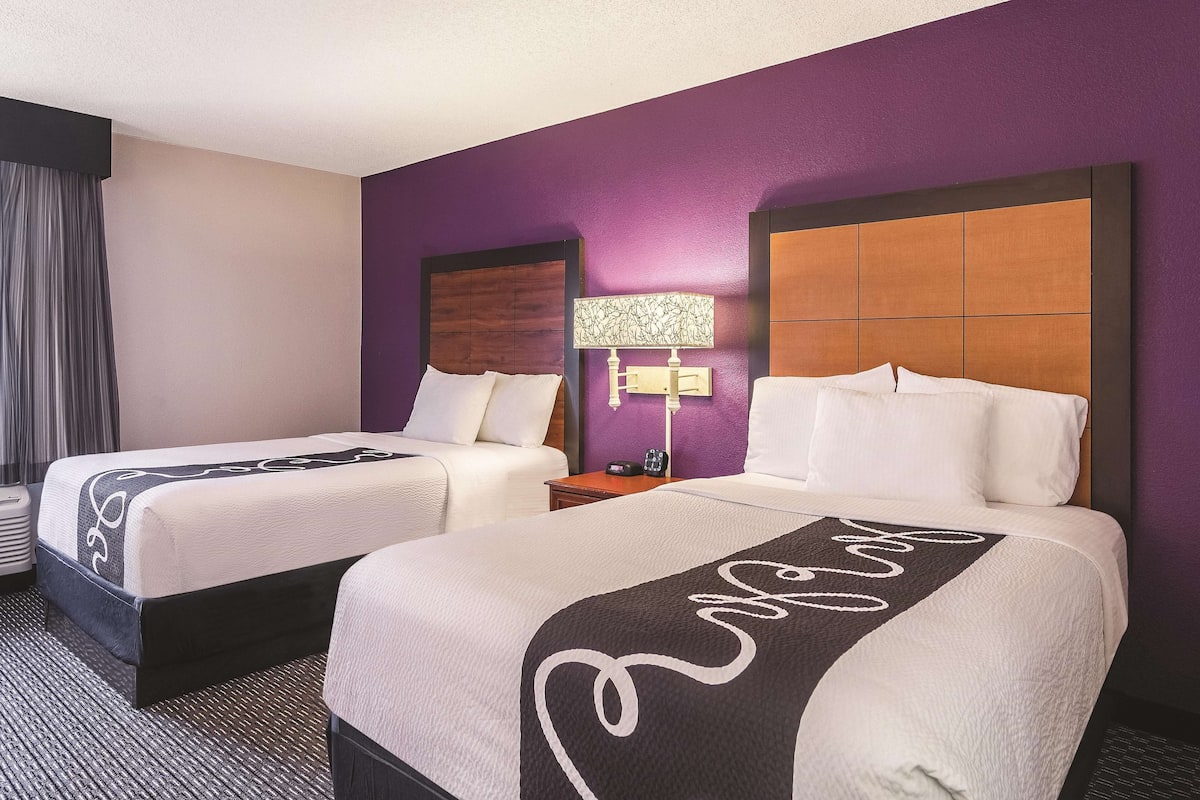 A hotel room with two beds, a nightstand, a lamp, and a window with curtains.