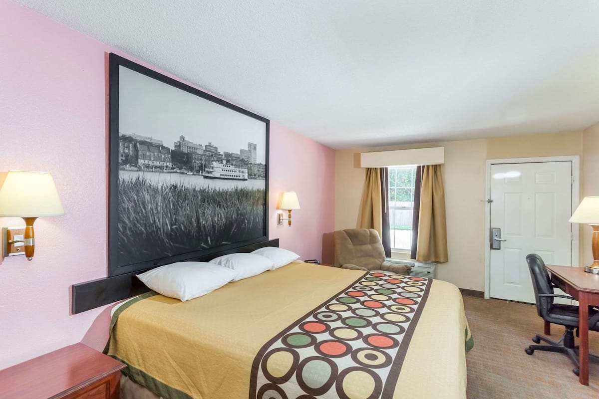 Een hotelkamer met een bed, een bureau, een stoel, een deur en een groot gepresenteerd schilderij aan de muur.