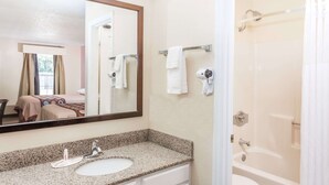 Une salle de bain d’hôtel avec un lavabo sur un comptoir en granit, un grand miroir, un espace douche et un lit visible dans la pièce adjacente.