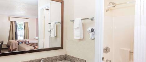 A hotel bathroom with a granite countertop sink, a large mirror, a shower area, and a visible bed in the adjacent room.