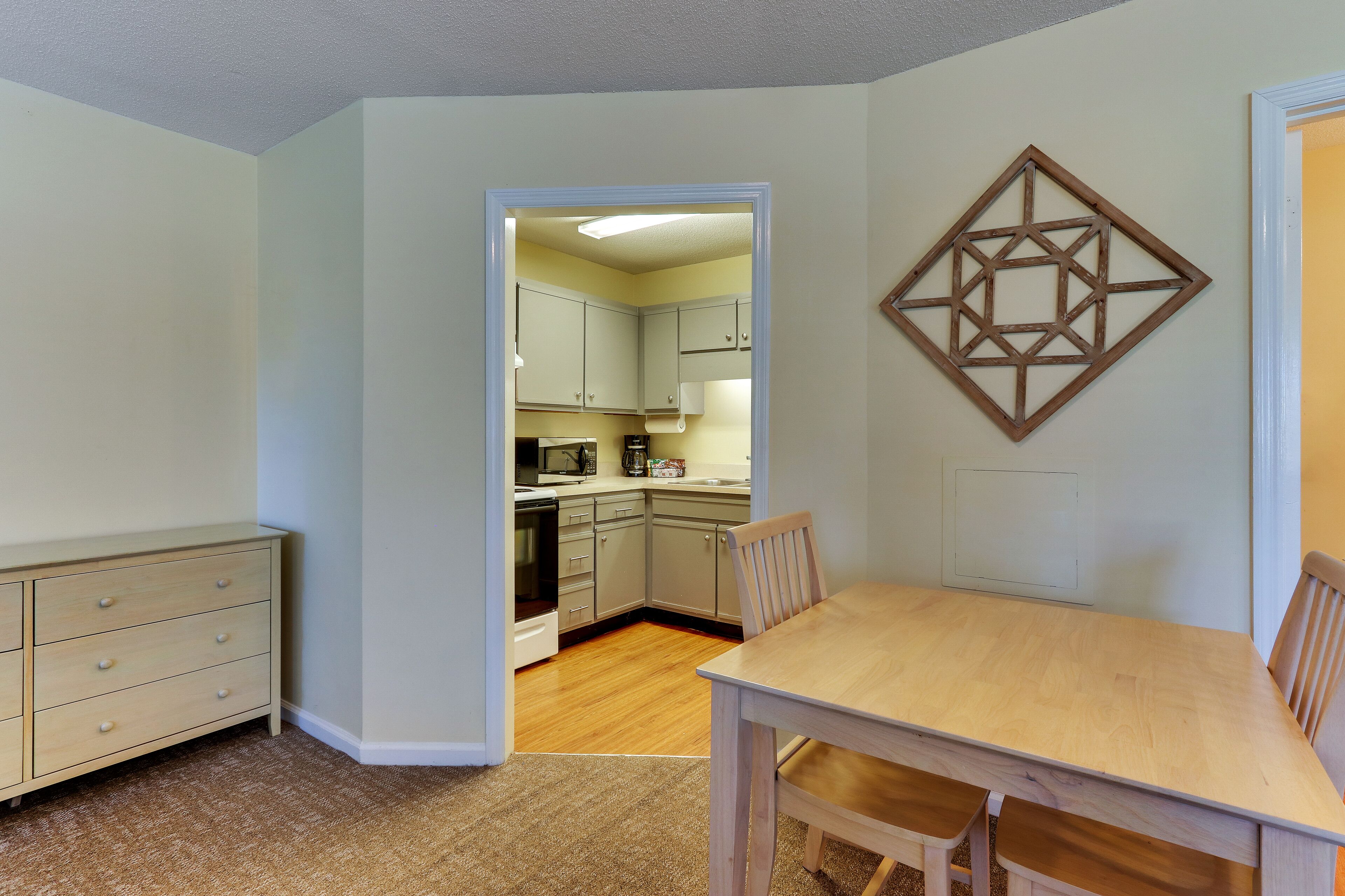 A compact kitchen with light-colored cabinets, a dining table with chairs, and a decorative geometric wall piece.