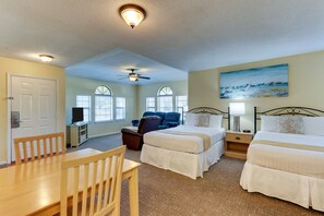 A hotel room with two beds, a desk, a chair, a ceiling fan, and a painting on the wall.