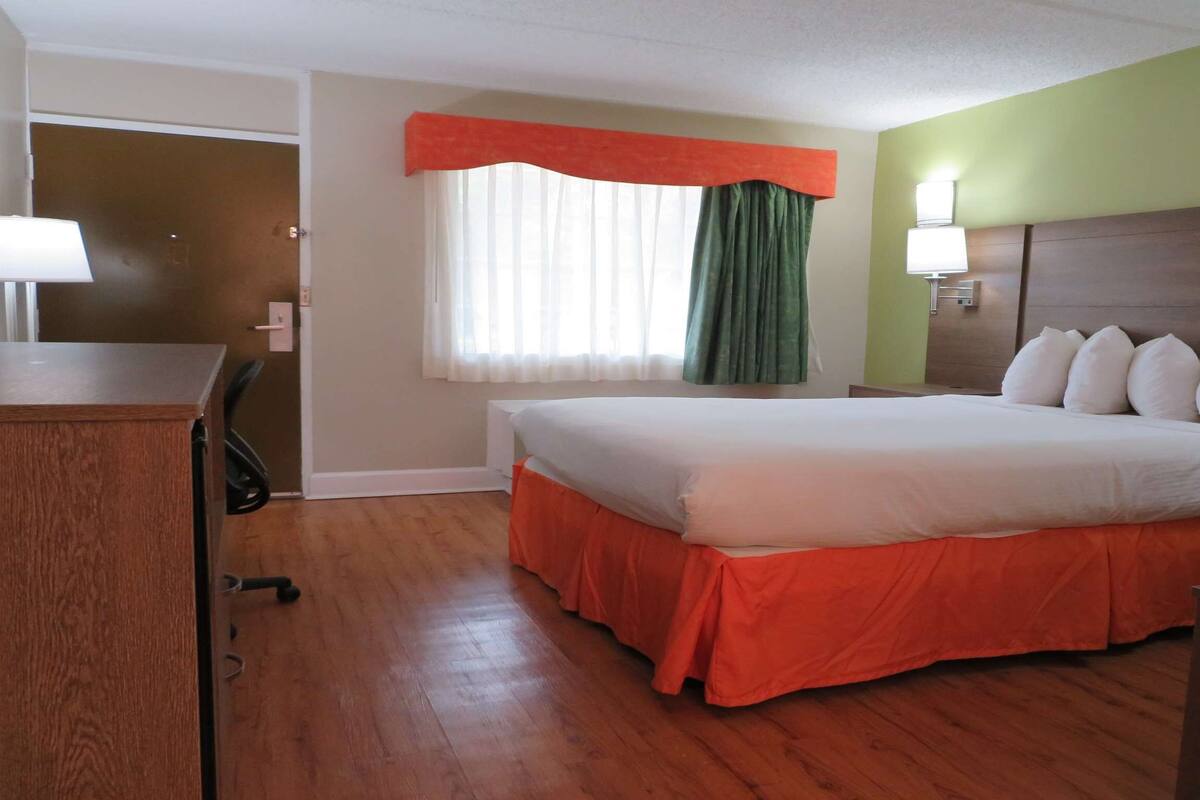A hotel room with a bed, a desk, a chair, a window with curtains, and a wall-mounted lamp.