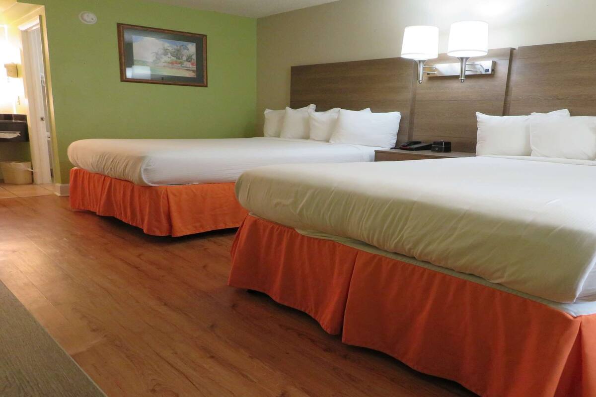 A hotel room with two beds, each with white bedding and orange skirts, a wooden headboard, and a wall-mounted lamp.