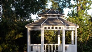 A white gazebo with a hexagonal shape and a peaked roof, surrounded by lush greenery.