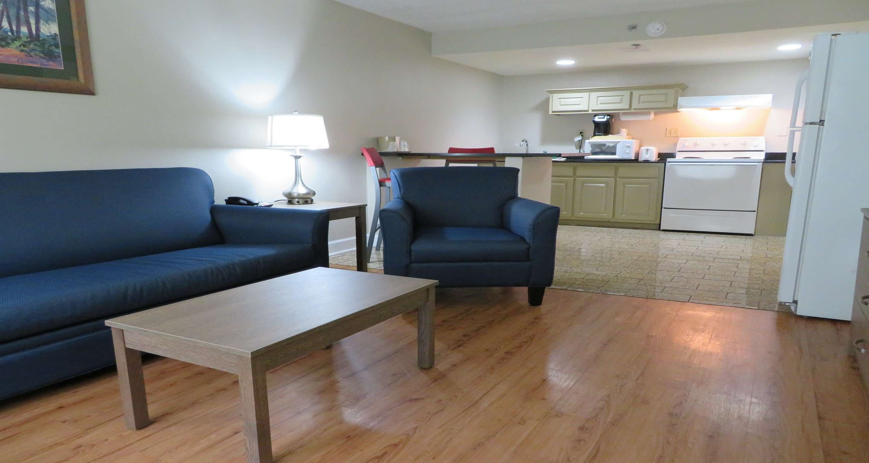 A living room with a blue sofa, a wooden coffee table, and a blue armchair. A kitchen with a refrigerator, stove, and microwave is visible in the background.