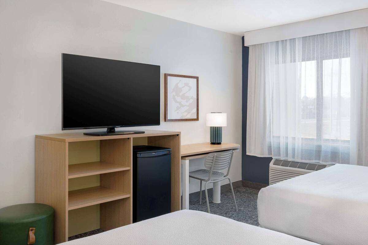 A hotel room with a flat-screen TV, a desk with a lamp, a chair, a bed, and a window with sheer curtains.