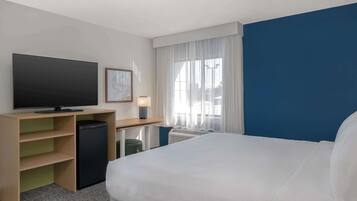 A hotel room with a bed, a desk with a television, a small refrigerator, a lamp, and a window with curtains.