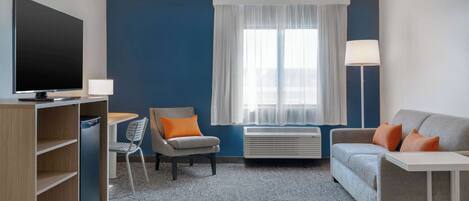 A hotel room with a flat-screen TV, a desk with a chair, a sofa, a standing lamp, and a large window with sheer curtains.