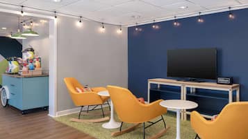 A modern room with a flat-screen TV, orange rocking chairs, a small table, and a kitchenette with a cart.