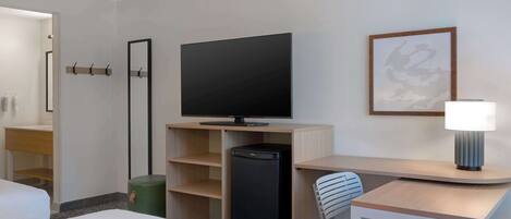 Habitación de hotel con cama, escritorio, silla, televisor, cesto de basura y lámpara de pared.