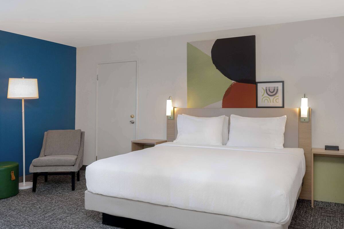 A hotel room with a large bed, a chair, a lamp, and a wall with abstract art.