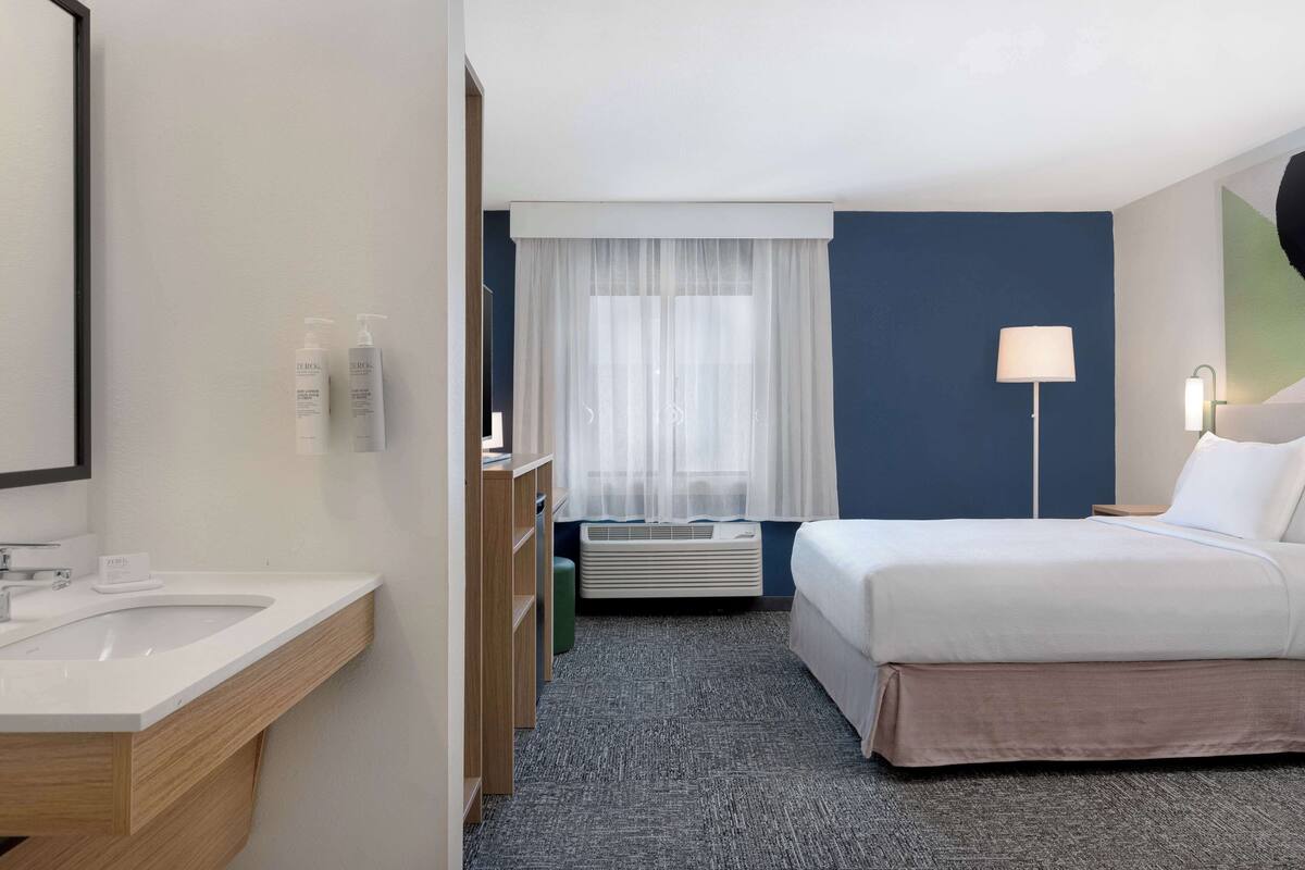 A hotel room with a bed, a bedside table, a lamp, a window with sheer curtains, a bathroom with a sink and toiletries, and a carpeted floor.