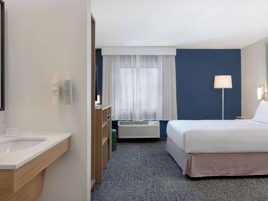 A hotel room with a bed, a bedside table, a lamp, a window with sheer curtains, a bathroom with a sink and toiletries, and a carpeted floor.
