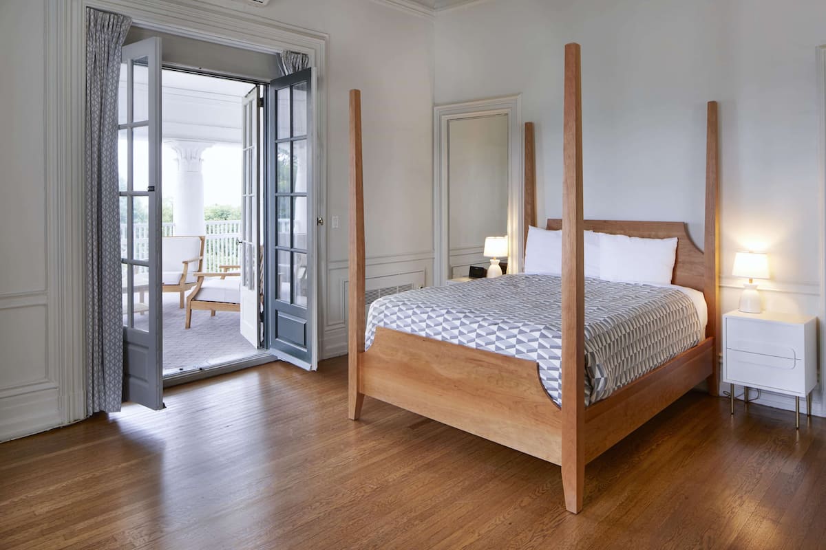 Een slaapkamer met een hemelbed, een houten vloer, een witte nachtkastje en uitzicht door een open deur op een buitenbalkon.