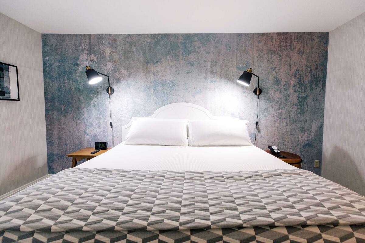 Een net opgemaakt bed met een bedsprei met een geometrisch patroon, twee bedlampjes, een nachtkastje met een telefoon, en een gestructureerde muur achter het bed.
