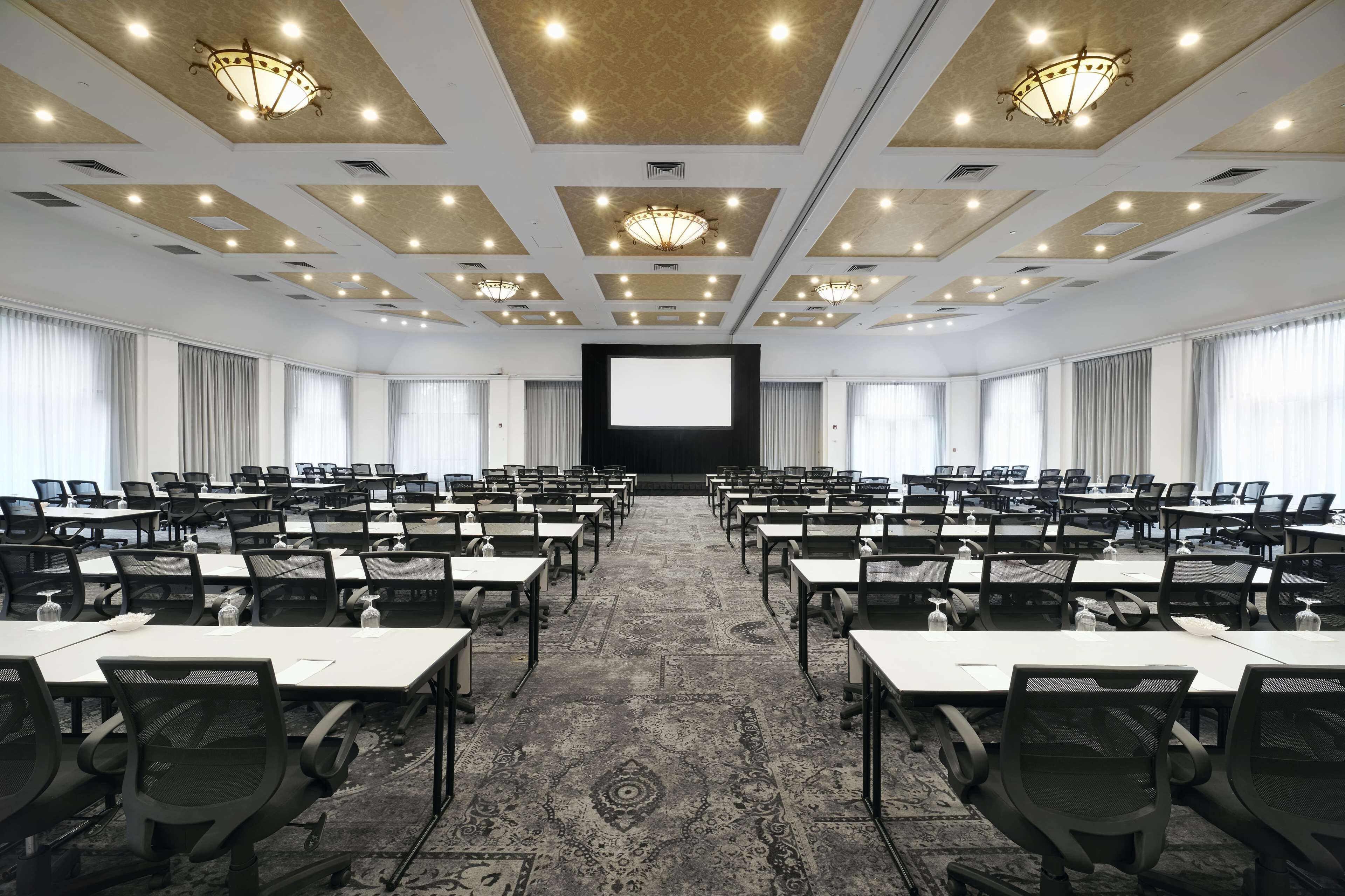 Un salón de conferencias con filas de mesas y sillas, una pantalla grande al frente y luces en el techo.