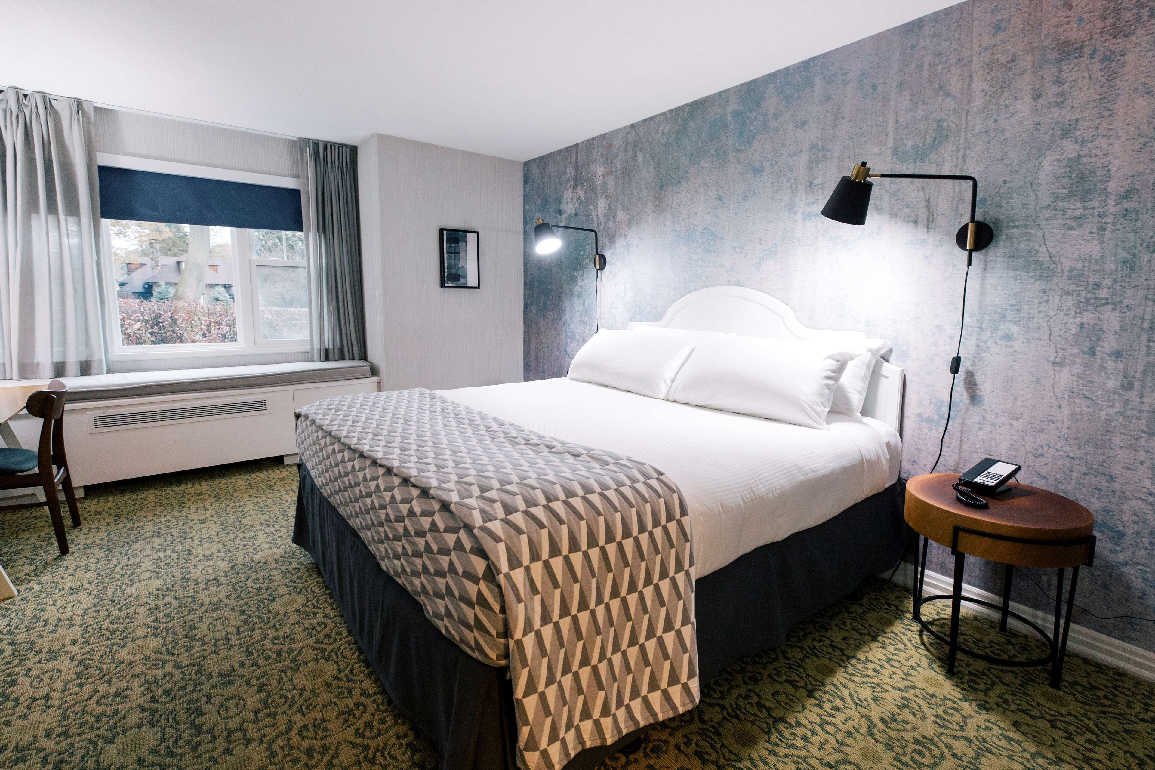 A neatly arranged hotel room with a bed, a desk, a chair, a round side table, and a lamp.