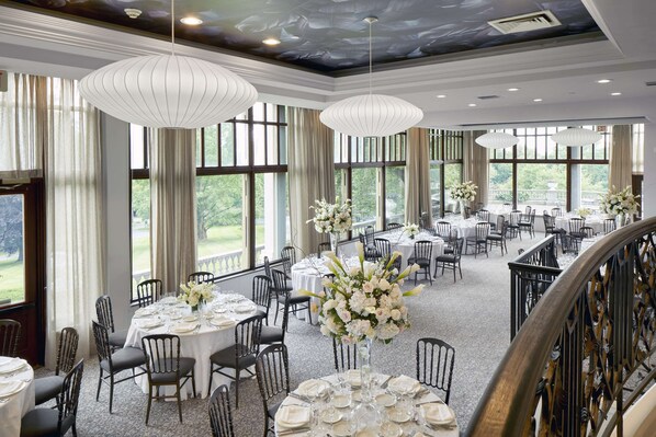 Un amplio salón de banquetes con varias mesas redondas dispuestas para comer, ventanas grandes que ofrecen vistas al exterior y elegantes candelabros.