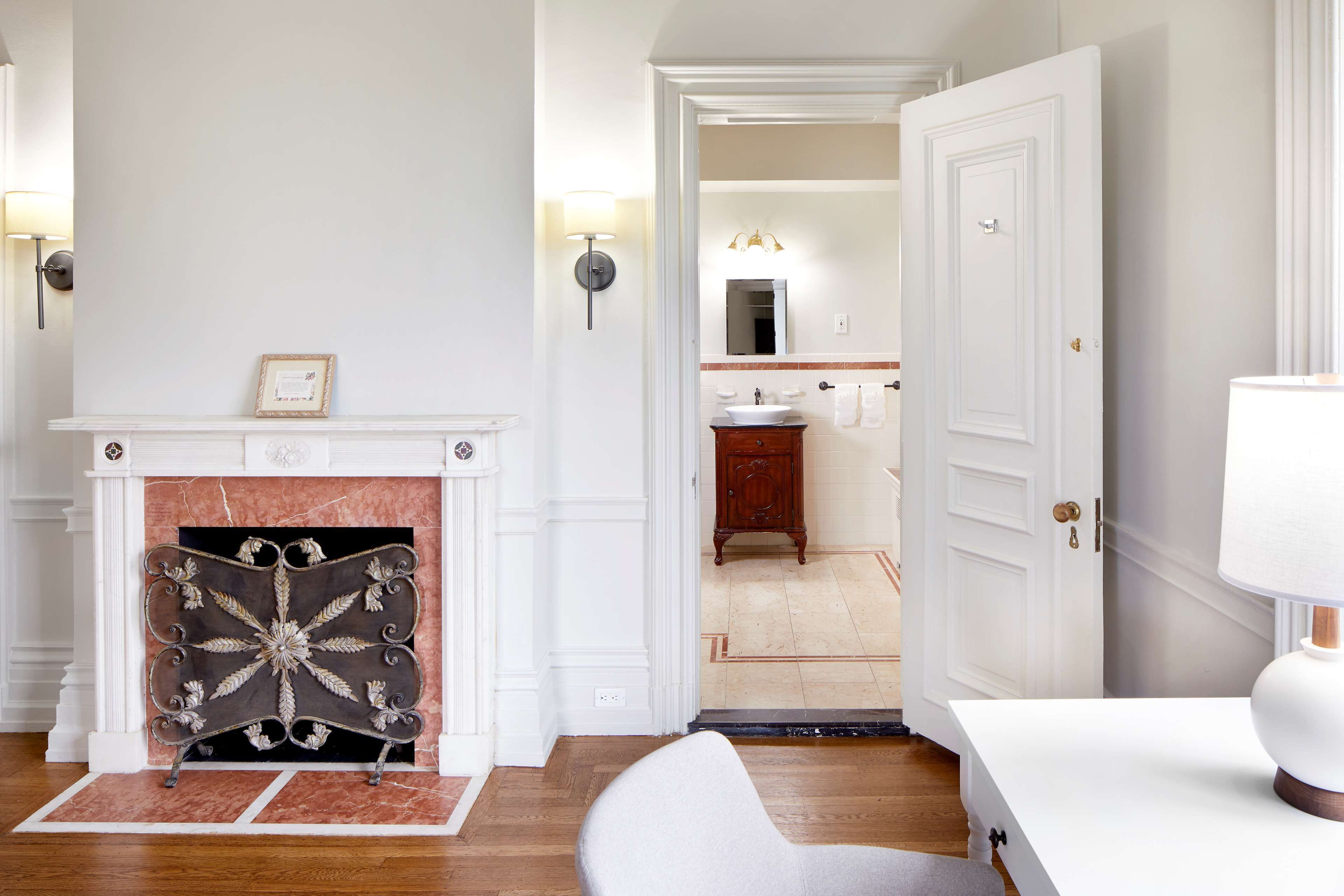 A room with a fireplace, a white wall, a wooden floor, a white door, and a bathroom visible through an open door.