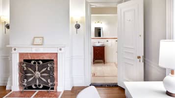 Ein Zimmer mit Kamin, weißer Wand, Holzboden, weißer Tür und einem durchgehenden Türspalt sichtbaren Badezimmer.